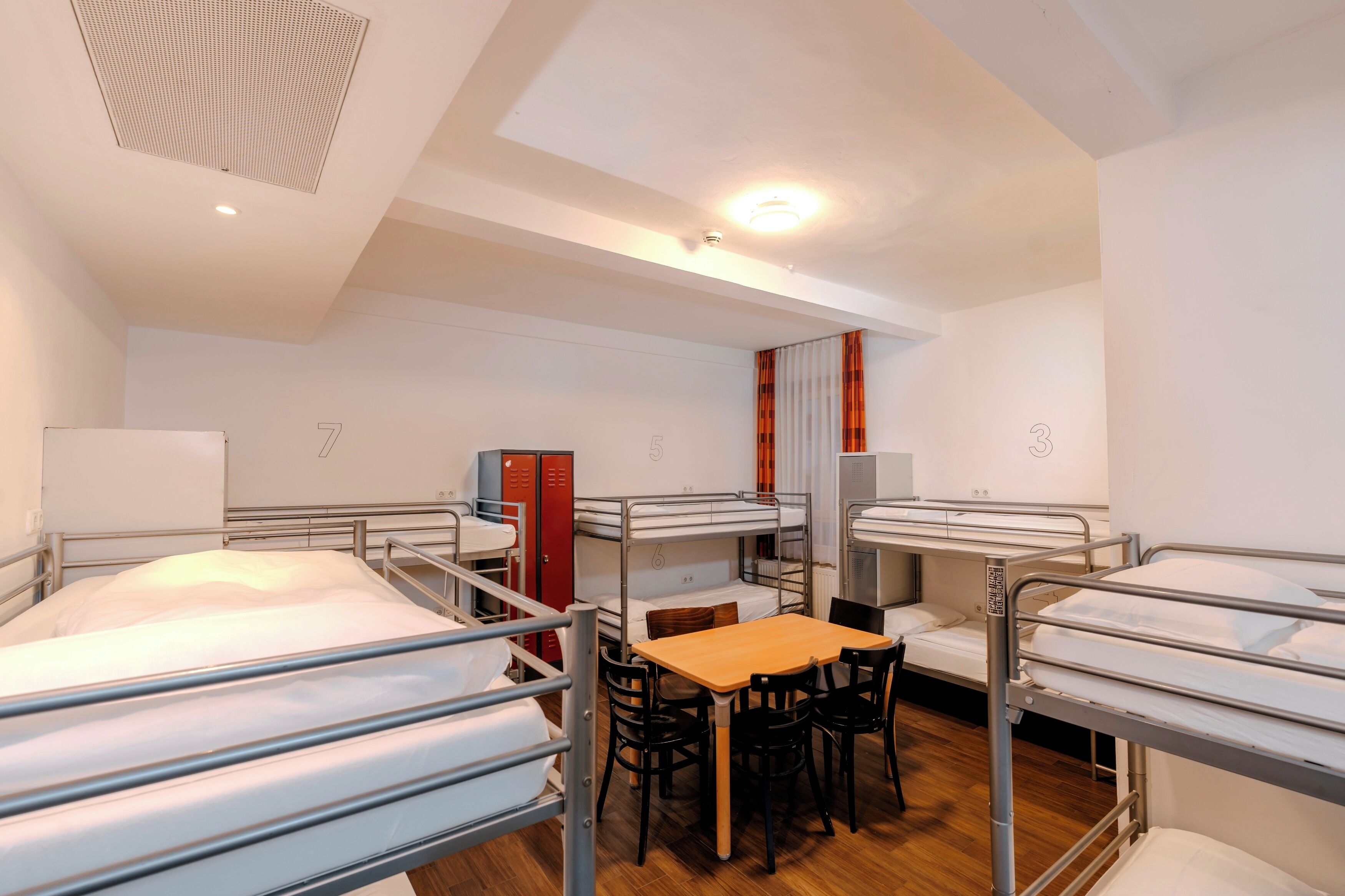 A dormitory room with bunk beds, a wooden table, chairs, and a desk.