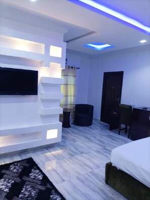 A modern bedroom with a flat-screen TV mounted on a white wall, a bed with a white headboard, a desk with a chair, and a small round table.