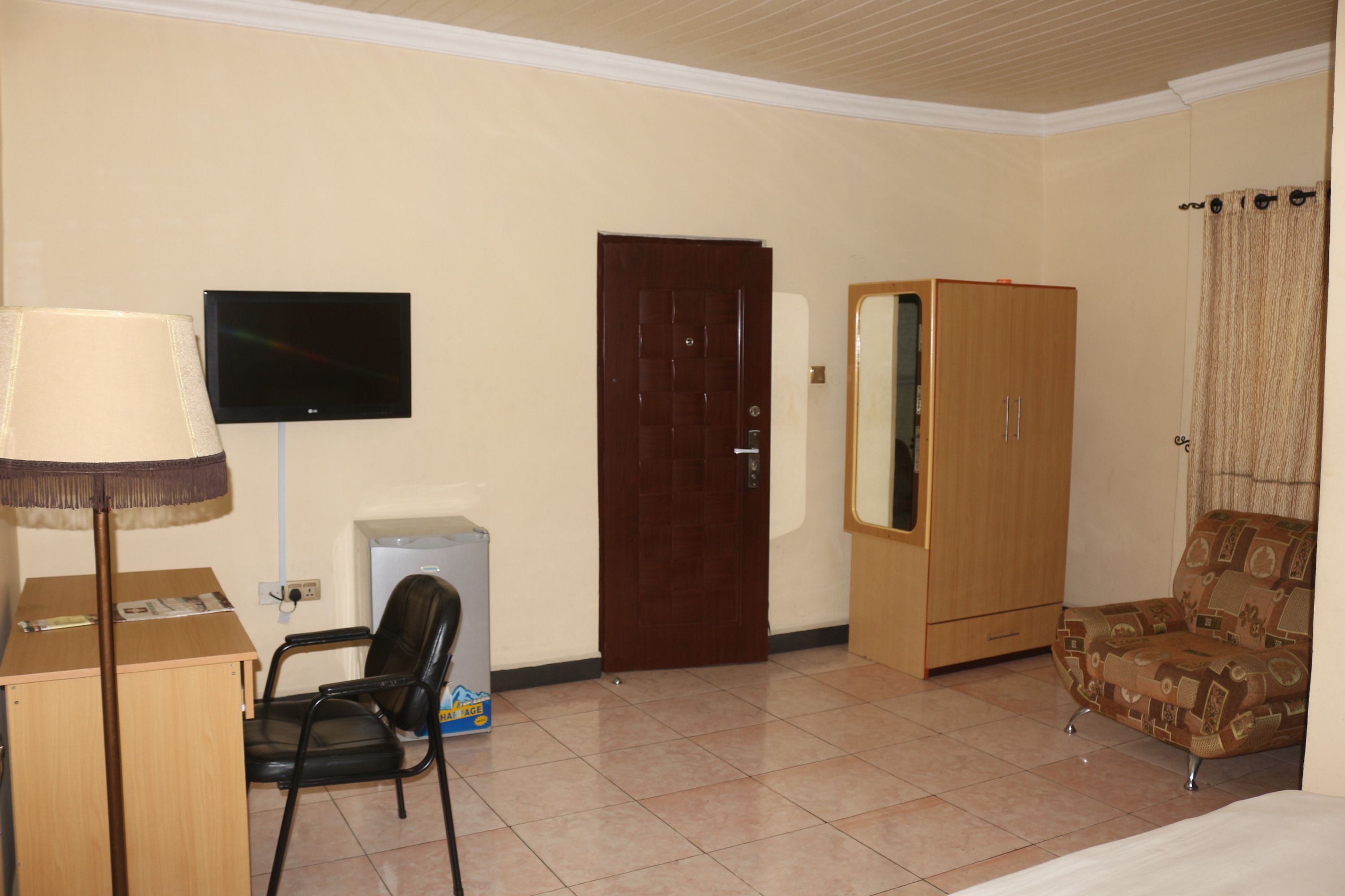 A hotel room with a bed, a desk with a lamp, a TV, a wardrobe, a chair, and a door.