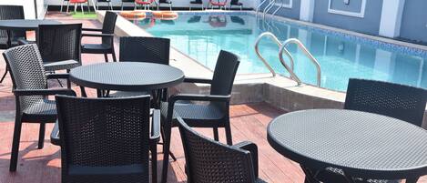 Uma área à beira da piscina com várias mesas redondas e cadeiras, uma piscina e um guarda-corpo.
