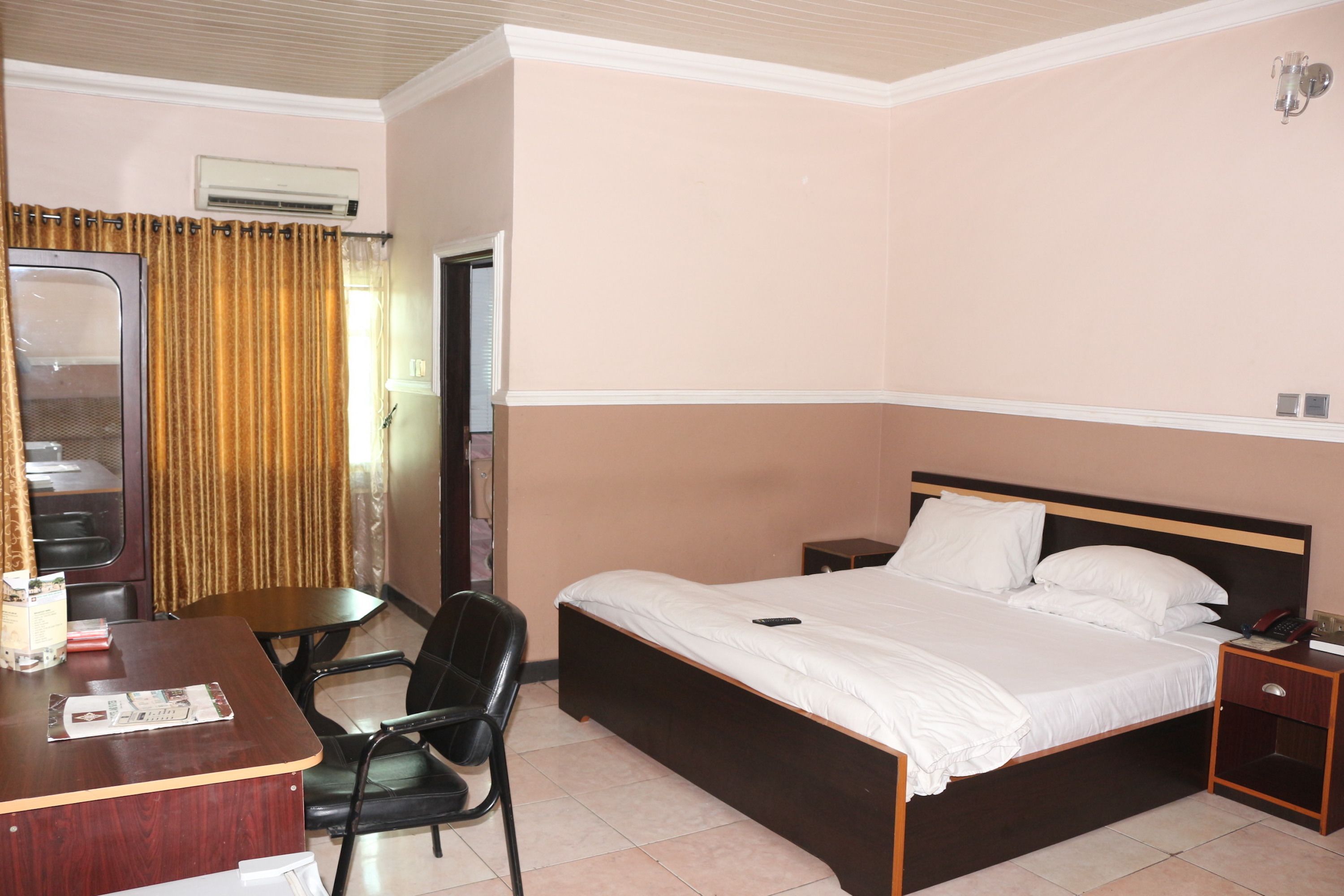 A hotel room with a bed, bedside table, chair, desk, and an air conditioning unit.