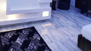 A modern living room with a flat-screen TV mounted on a white wall, a black ottoman, and a patterned rug. - NEW POINT HOTEL AND SUITES (Uyo)