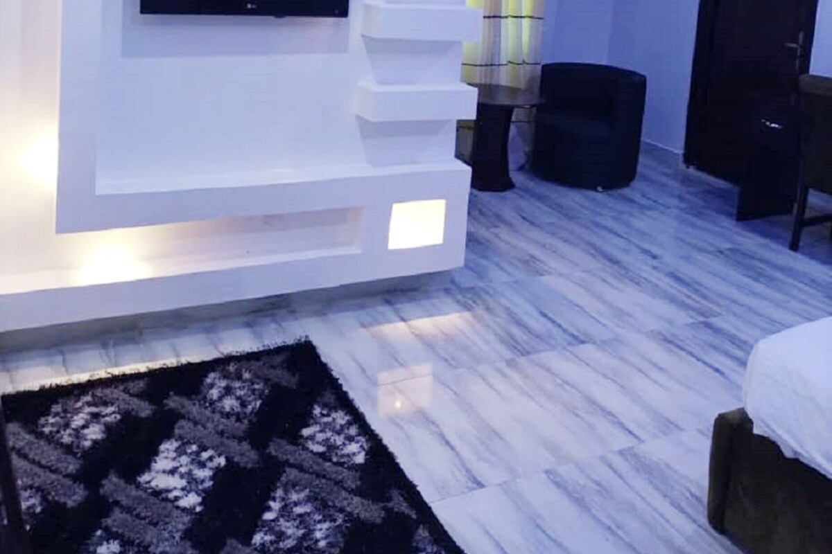 Een moderne woonkamer met een flatscreen televisie aan een witte muur, een zwart voetenbankje en een kamerbreed tapijt met een patroon.
