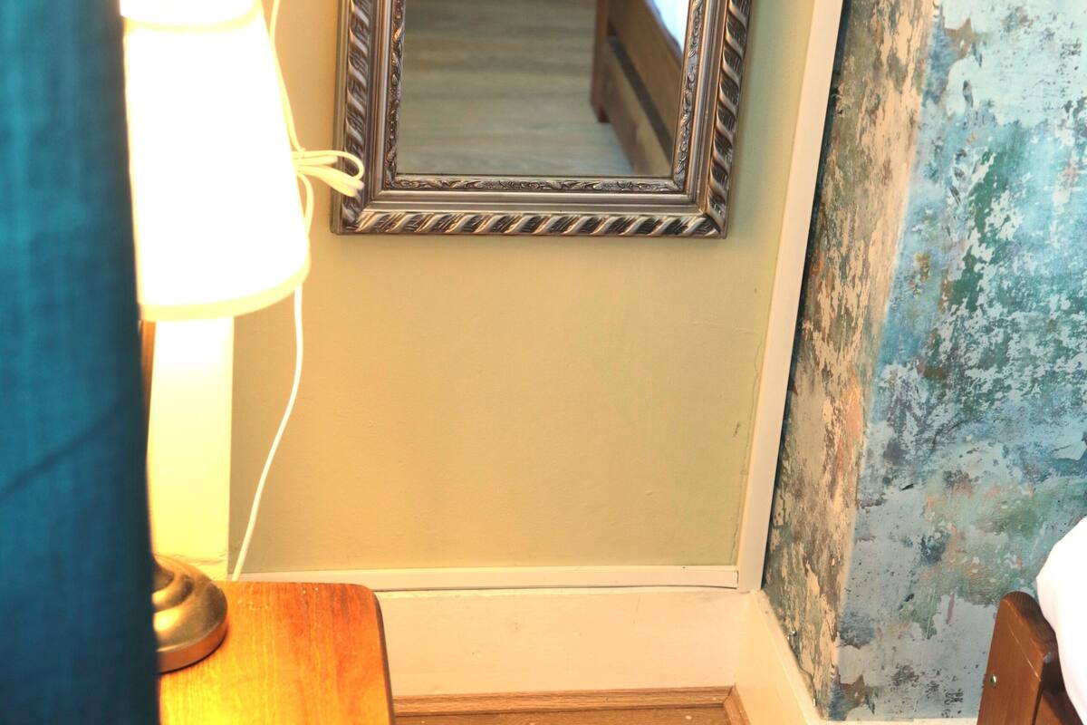 A hotel room with a wooden floor, a bed, a nightstand with a lamp, and a mirror reflecting a bed and a wall with peeling paint.