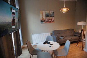 A modern living room with a grey sofa, a round white table, and two grey chairs. There is a flat-screen TV mounted on the wall, a radiator, and a floor lamp.