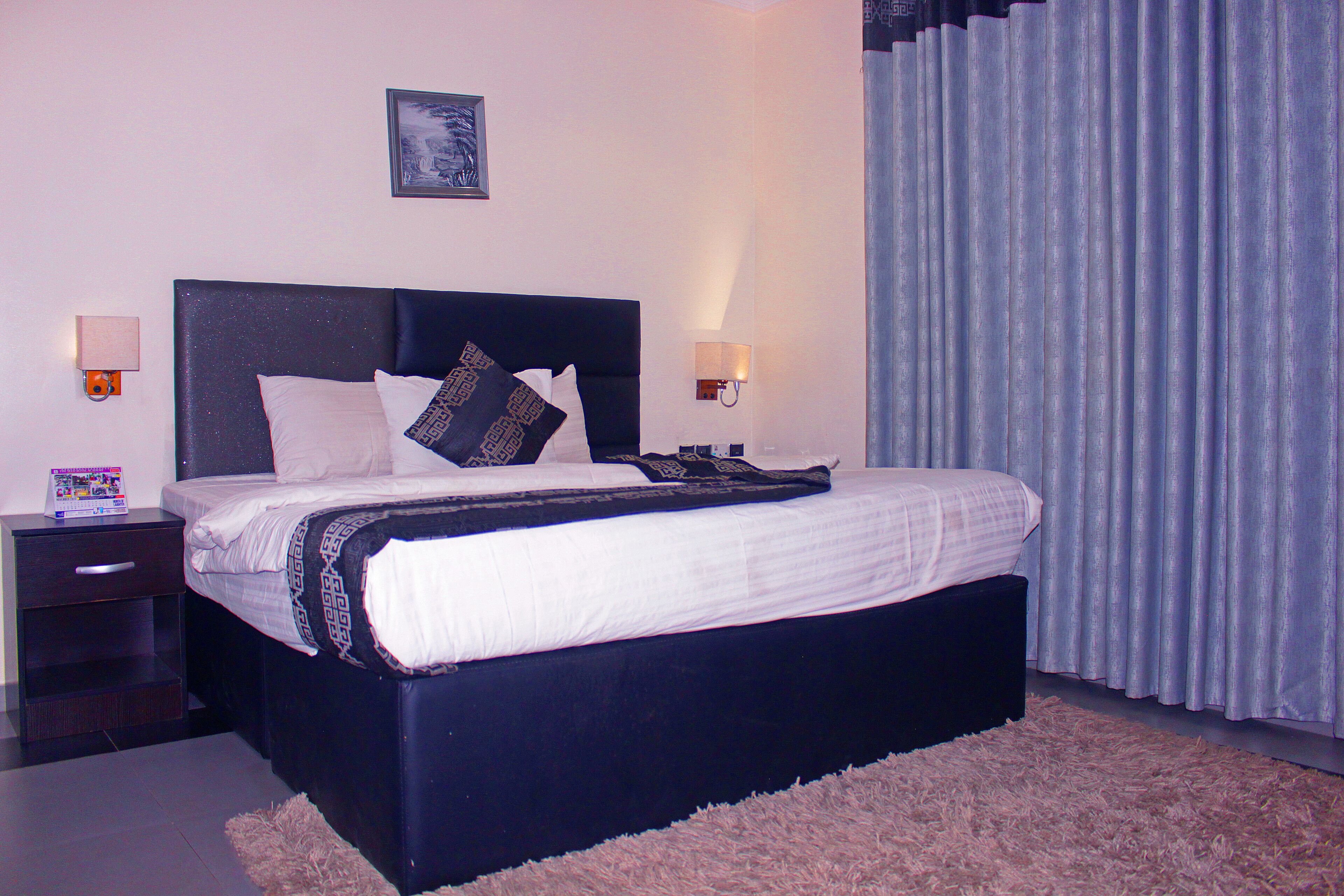 A neatly made bed with a dark headboard, a bedside table with a lamp, and a large window with curtains.
