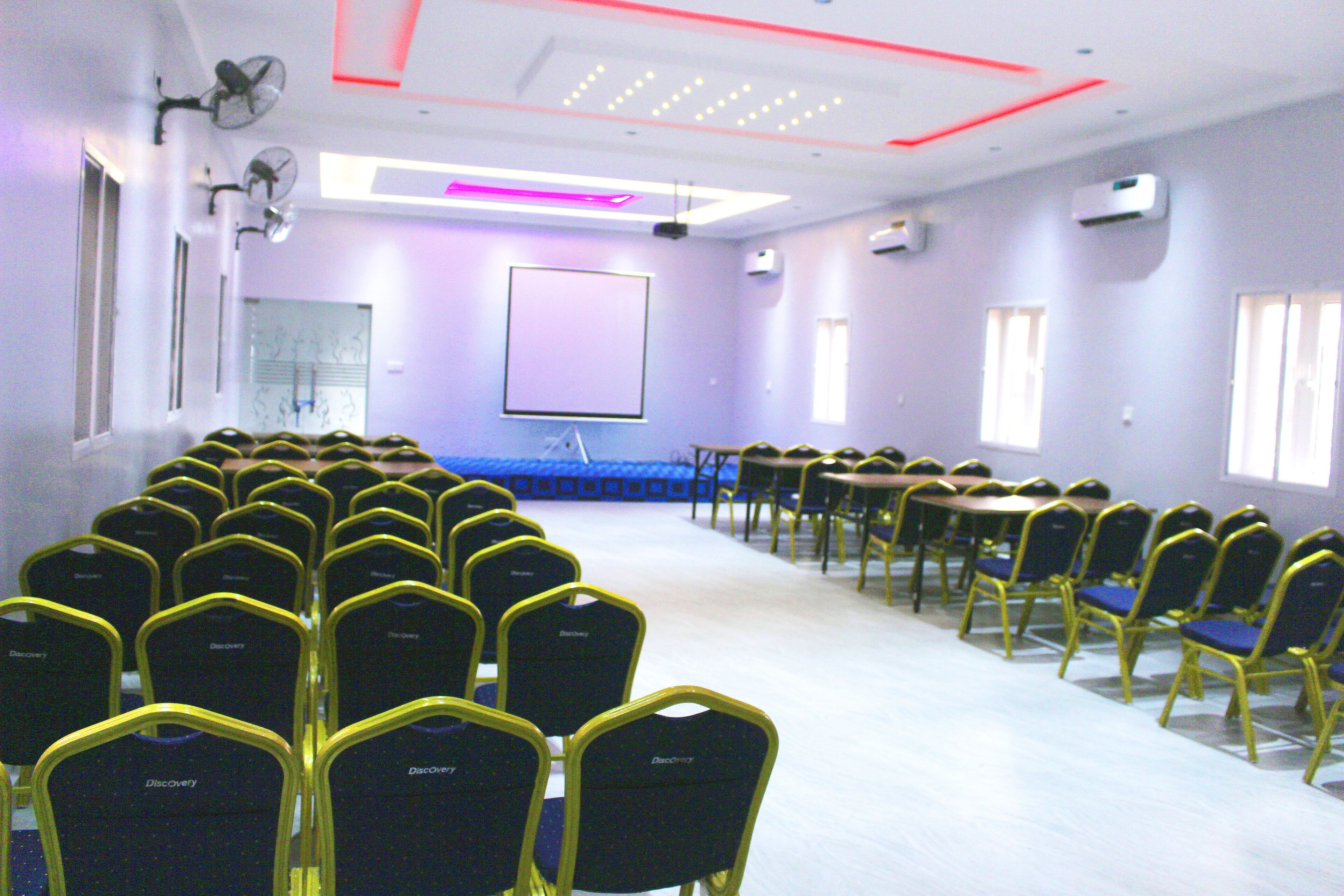 Une salle de conférence avec des rangées de chaises jaunes et bleues, un écran de projection et des ventilateurs de plafond.