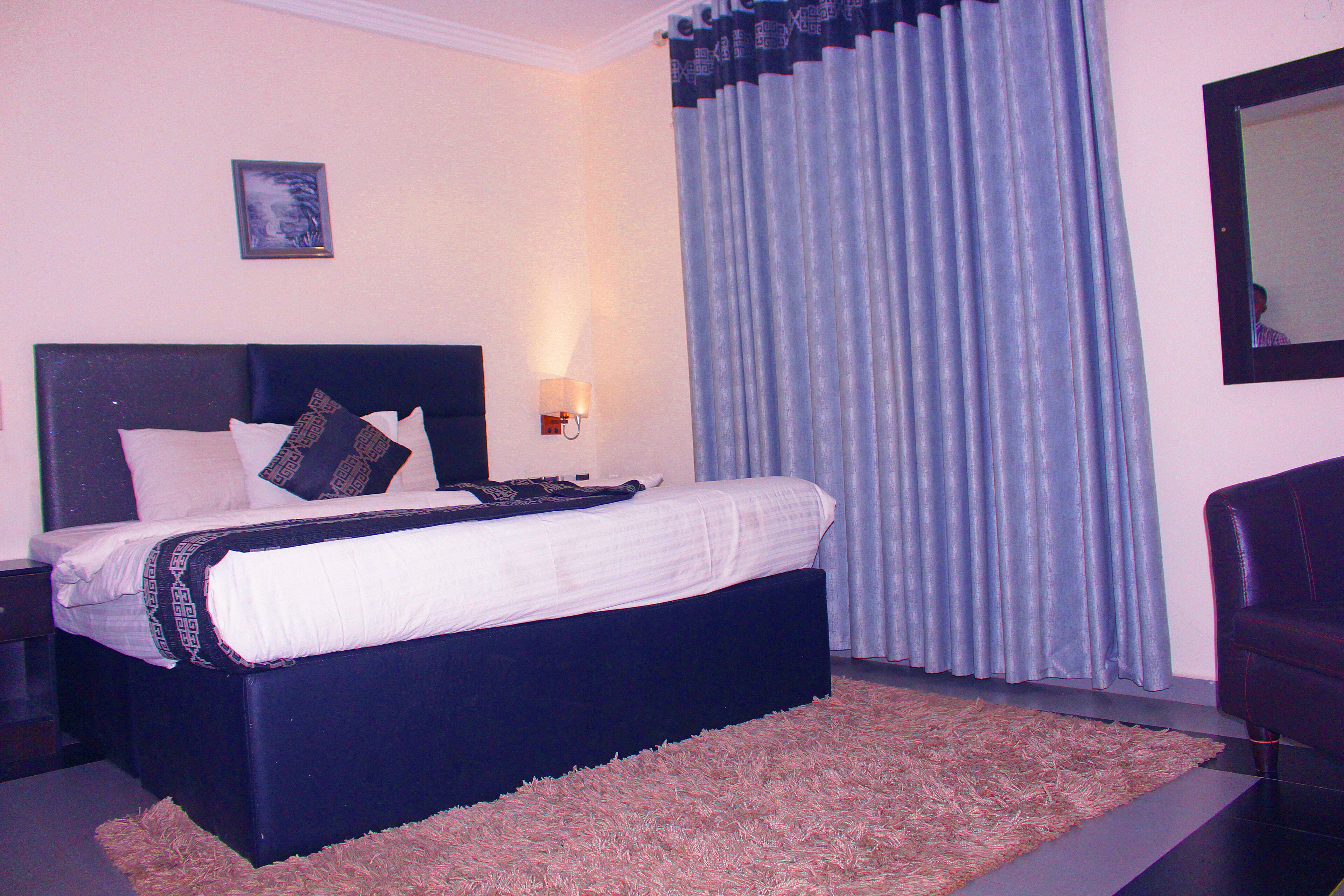 A bedroom with a bed, a nightstand, a lamp, a large window with curtains, and a mirror on the wall.