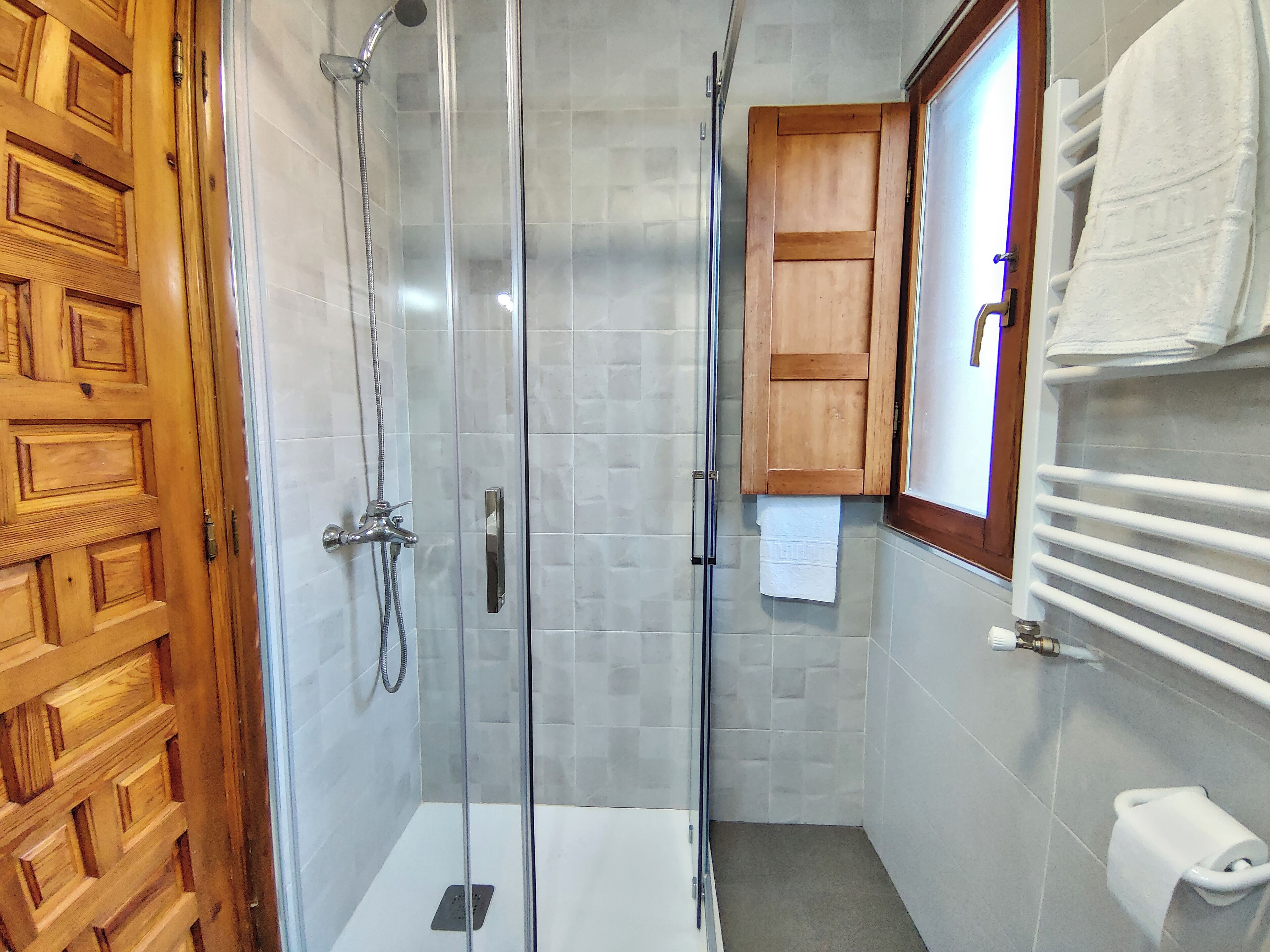 Une douche fermée par des parois de verre, dotée d’une porte en bois, de murs carrelés et d’un meuble en bois sur lequel est accrochée une serviette.