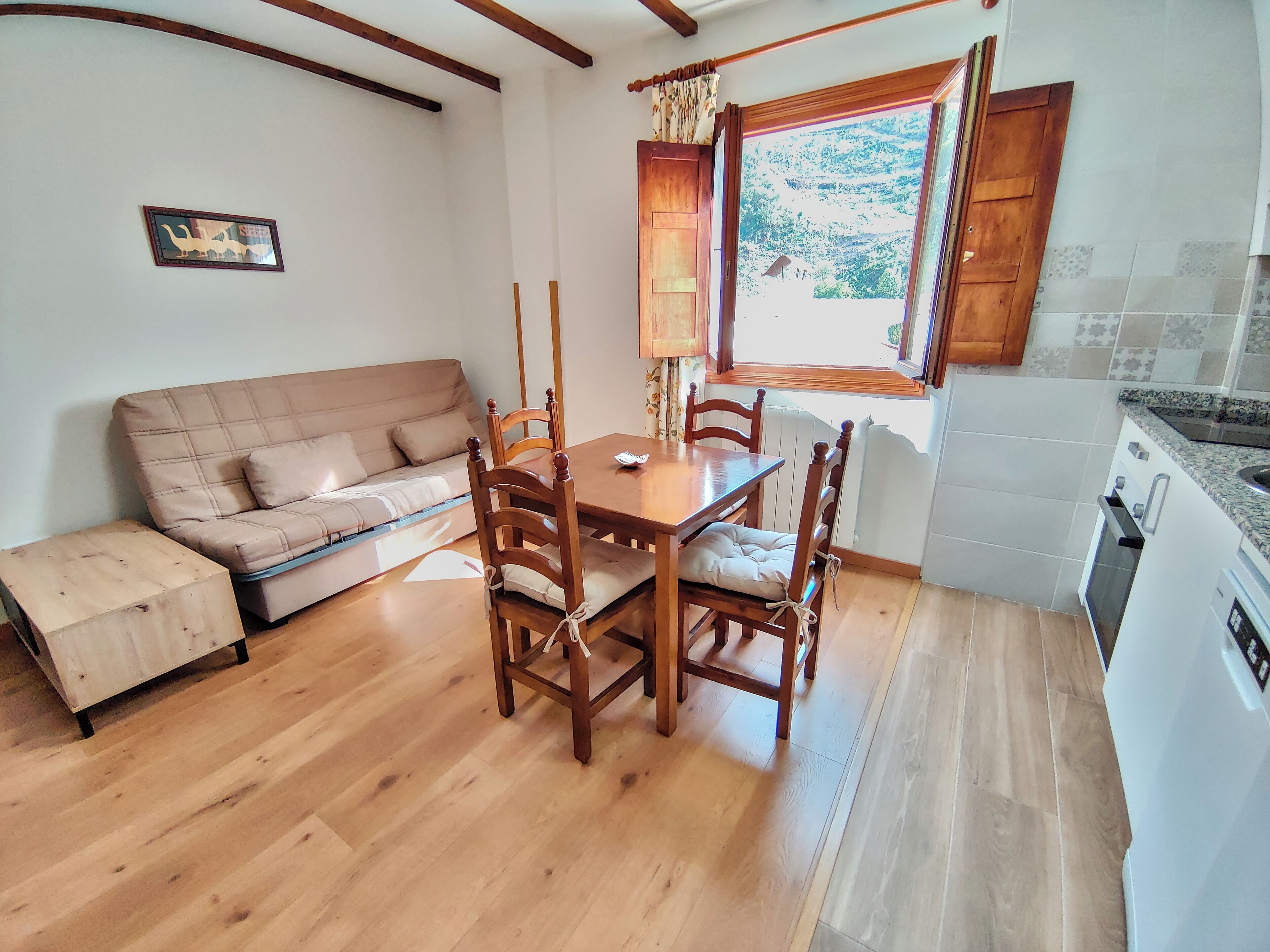 Una sala de estar acogedora con sofá, mesa de comedor y sillas, una zona de cocina con electrodomésticos, y una ventana con cortinas.