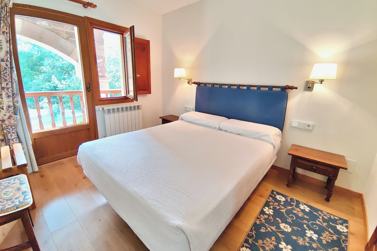 A bedroom with a large bed, two bedside tables, a window with a view, and a door leading to a balcony.