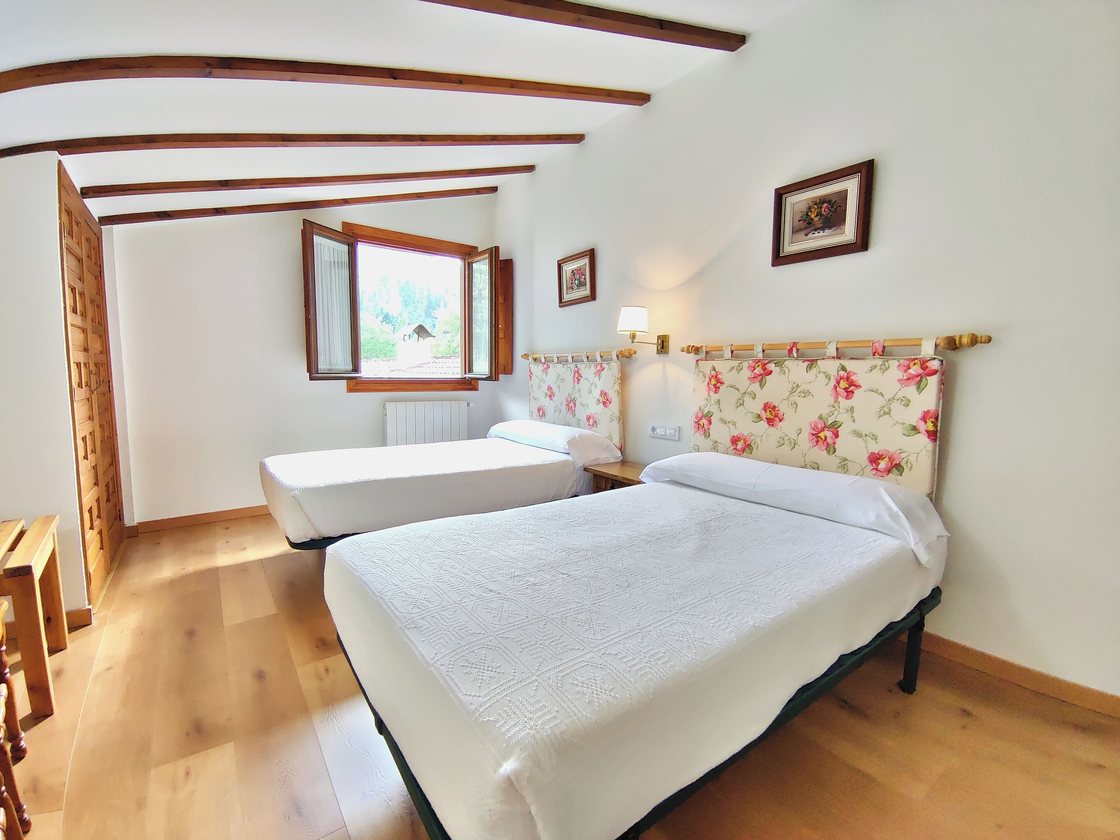 Un dormitorio con dos camas individuales, vigas de madera en el techo, una ventana con vistas a la vegetación y una lámpara fijada a la pared.