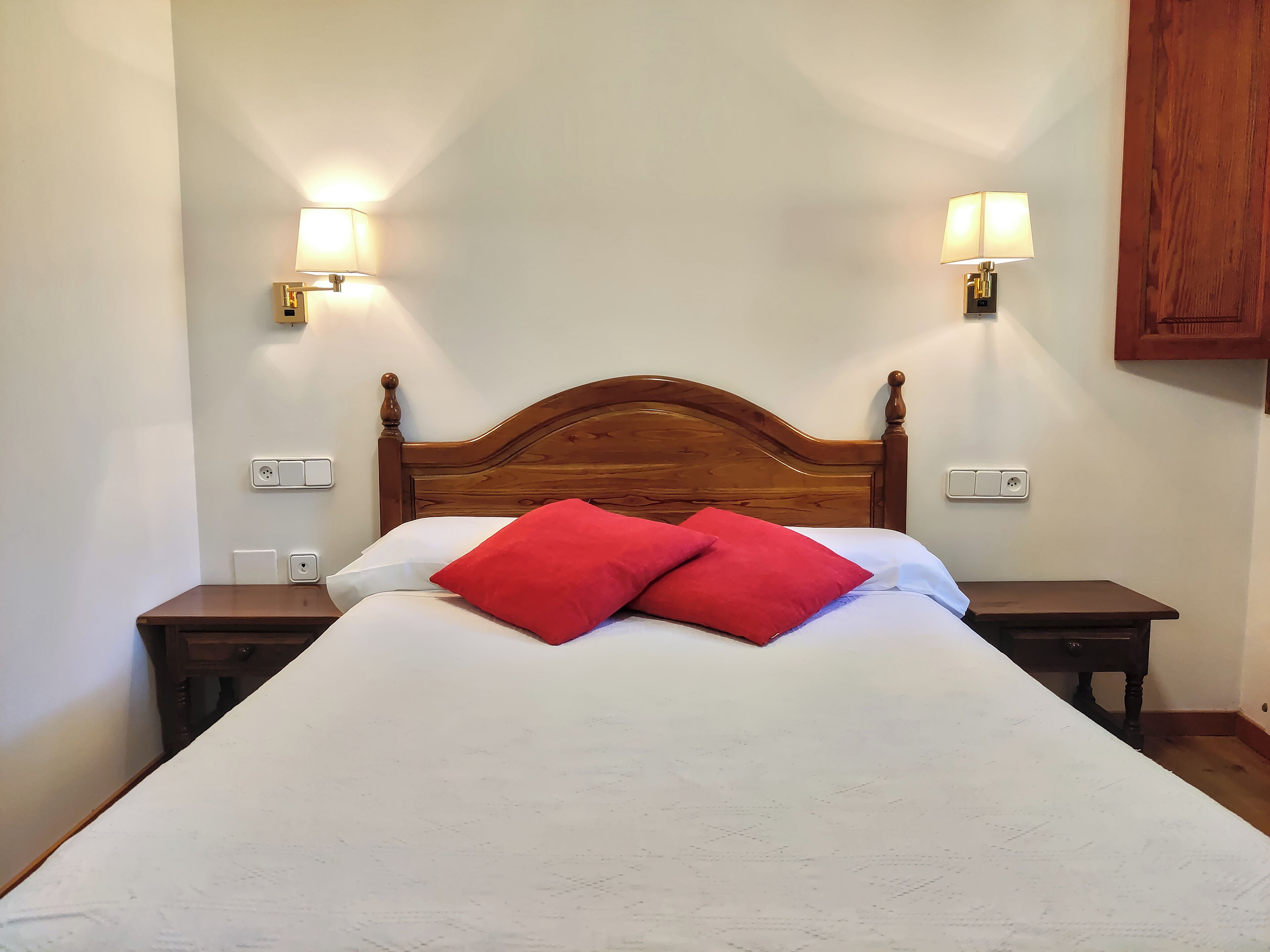 Una cama bien hecha con cabecera de madera, dos mesitas de noche y dos lámparas de pared.