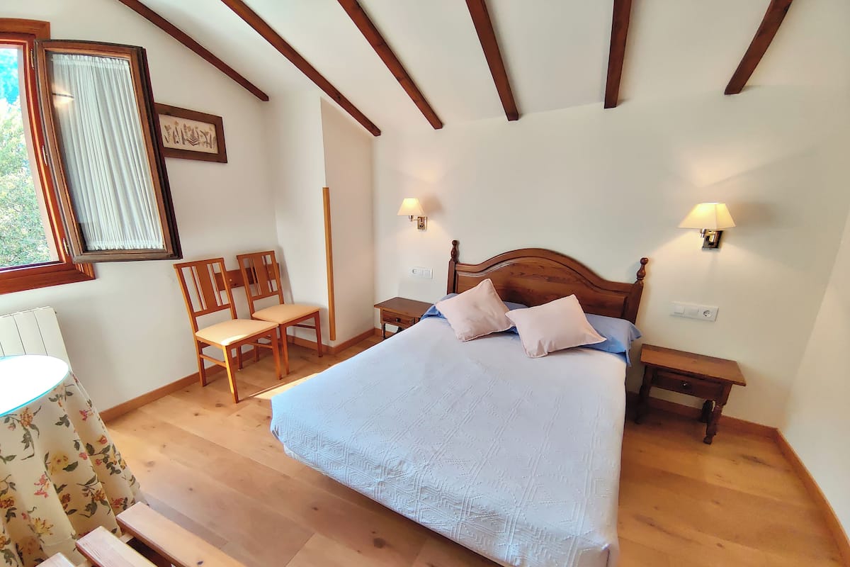A bedroom with a large bed, wooden headboard, two bedside tables with lamps, a wooden bench, a chair, and a window with curtains.