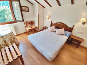 A bedroom with a wooden bed, two bedside tables, a nightstand, a chair, a table with a floral tablecloth, and a window with curtains. - Apartamentos Javier (Camaleno)