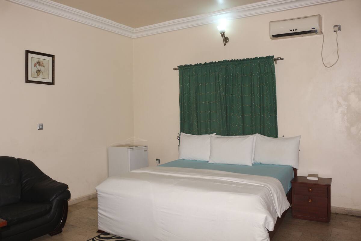 Quarto com cama, mesa de cabeceira, pequena mesa, cadeira e ar condicionado.