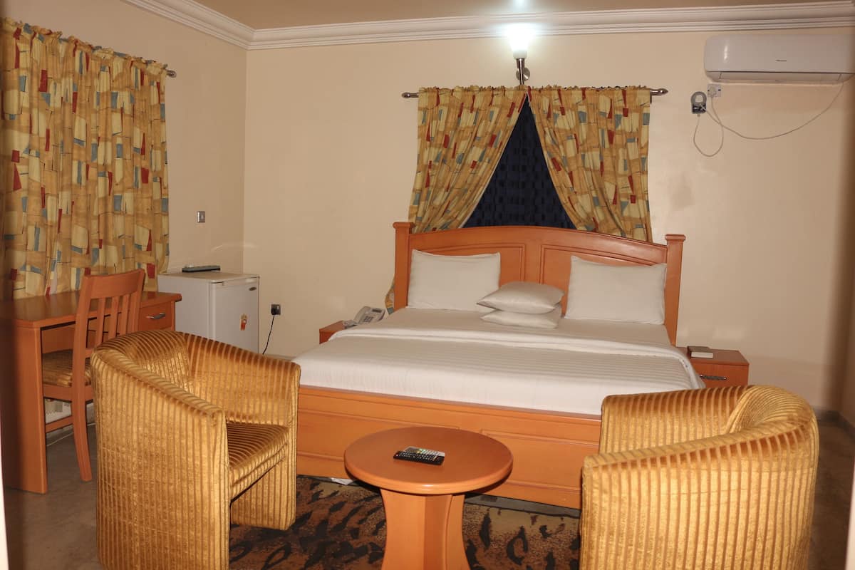 Quarto de hotel com cama de madeira, duas poltronas estofadas, uma mesa redonda pequena e uma escrivaninha com cadeira.