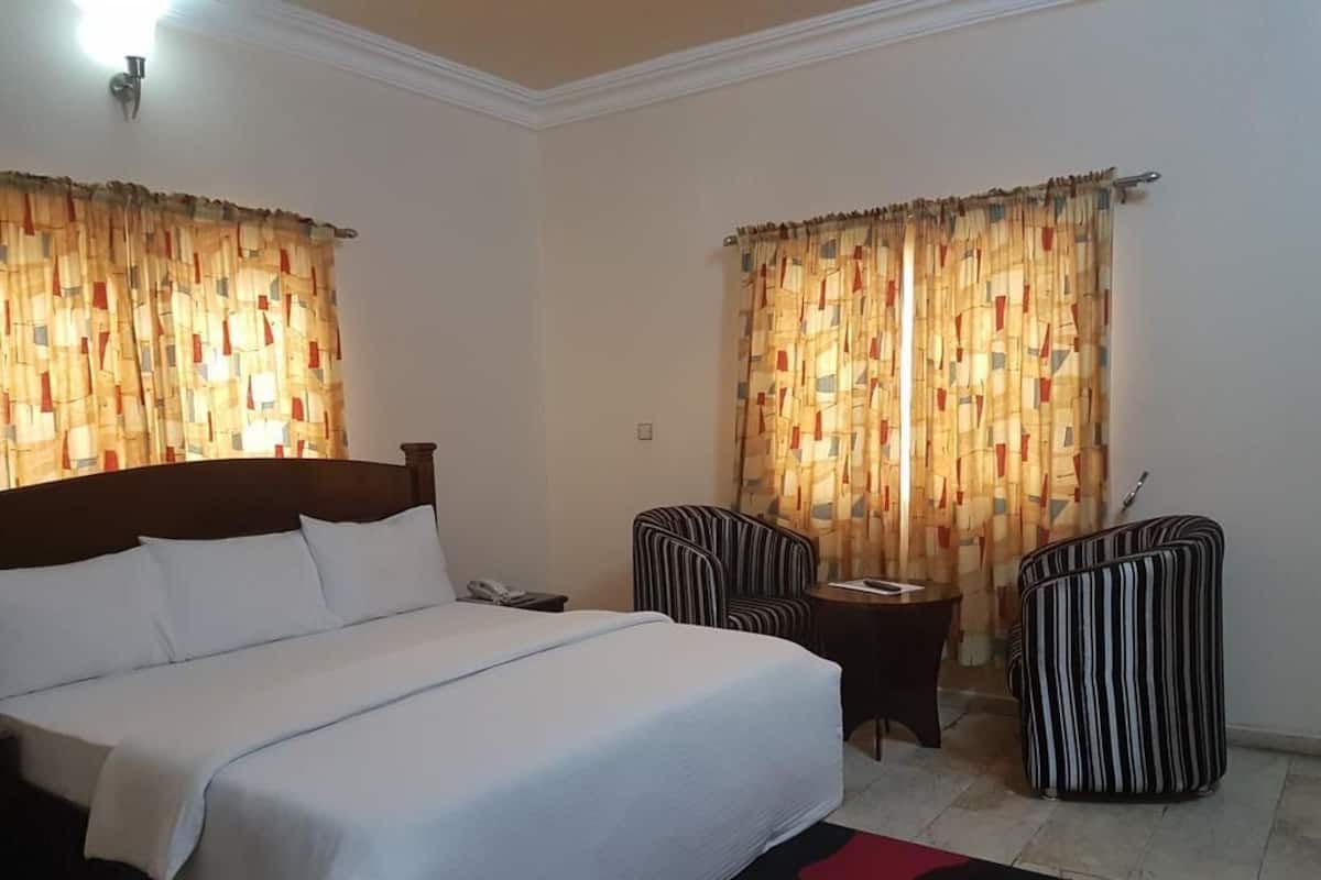 Quarto de hotel com cama, duas poltronas listradas, uma mesa pequena e cortinas.