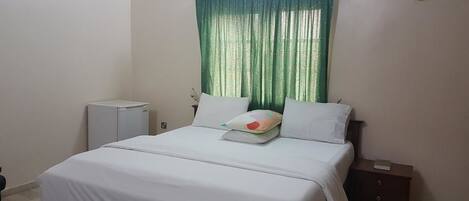 A bedroom with a bed, a nightstand, a small cabinet, a heater, and a window with green curtains.