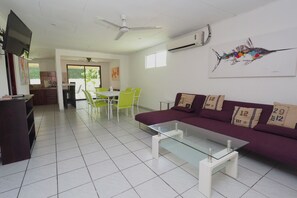 A modern living room with a glass coffee table, a purple sofa, and a dining area with green chairs.