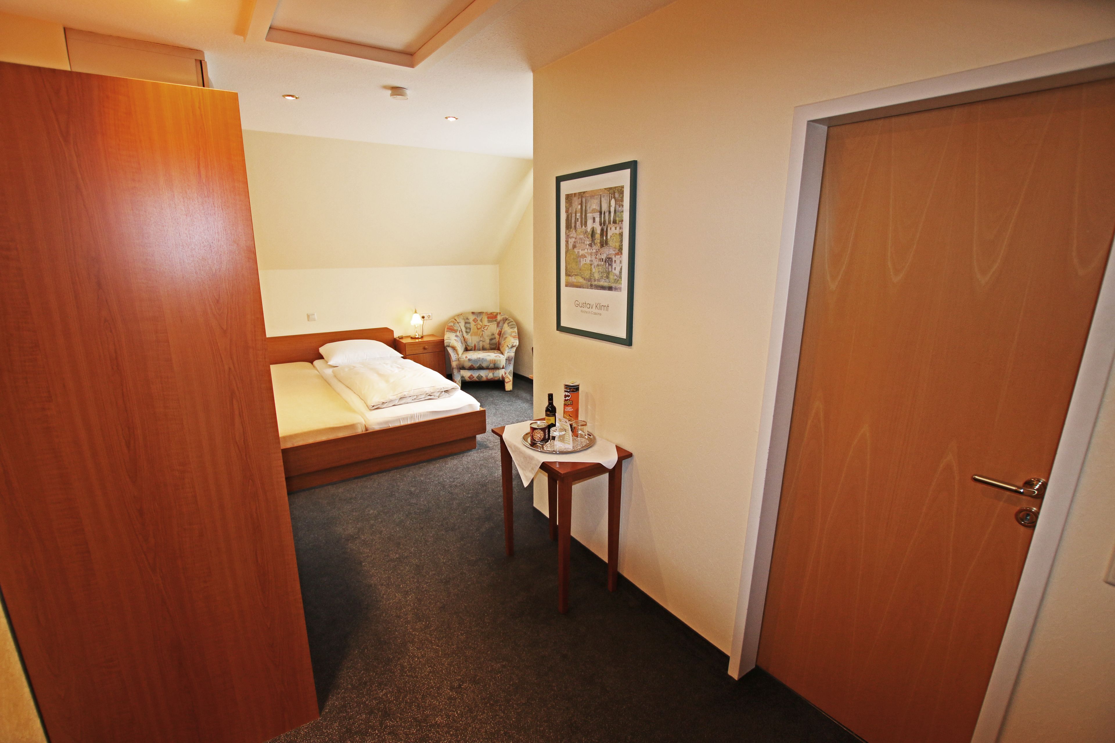 Habitación de hotel con cama, una mesita con una botella y vasos, una silla y una puerta.