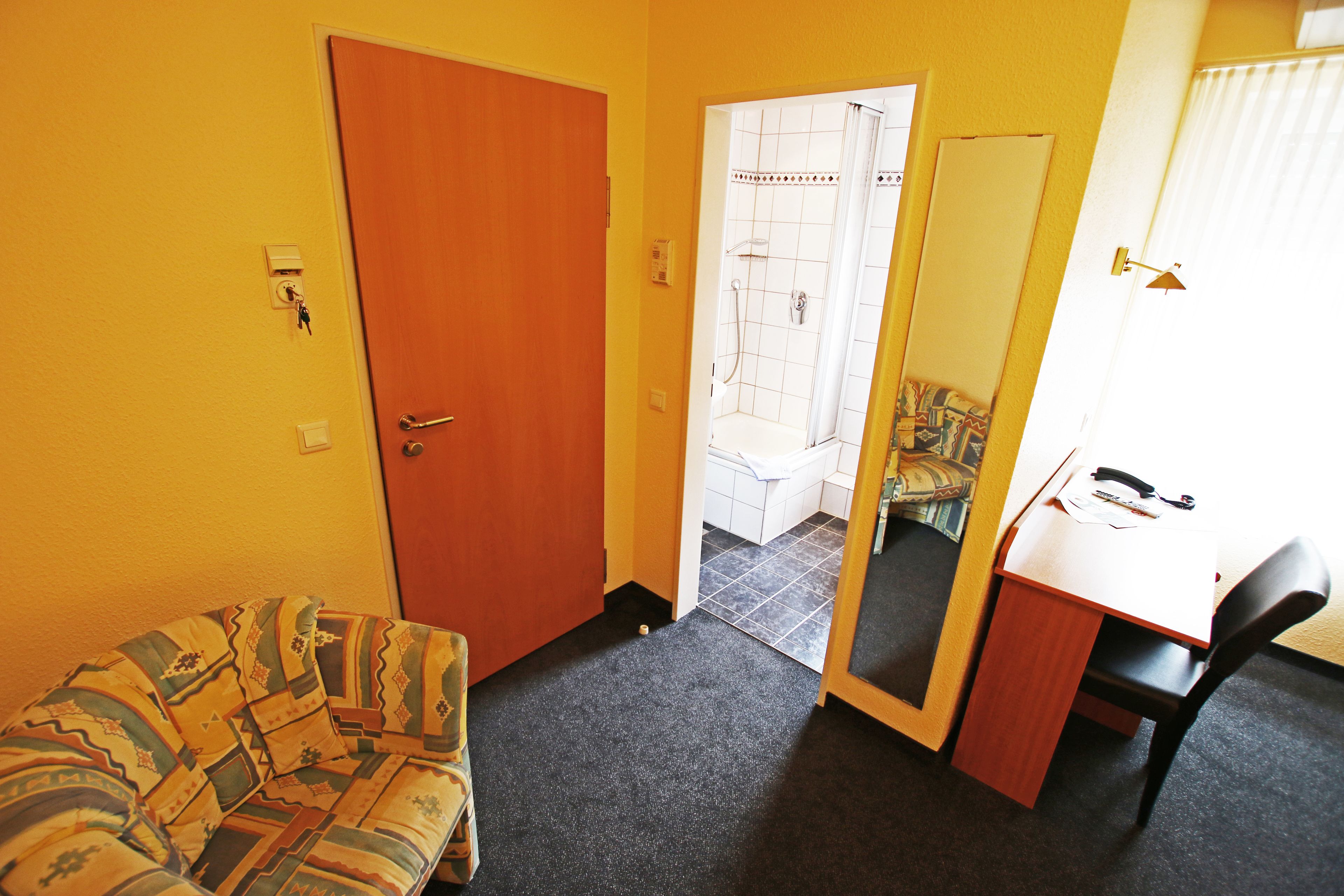 Een kleine kamer met een houten deur, een sofa met een patroon, een bureau met een stoel, en een douchegedeelte zichtbaar door een open deur.