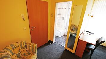 Een kleine kamer met een houten deur, een sofa met een patroon, een bureau met een stoel, en een douchegedeelte zichtbaar door een open deur.