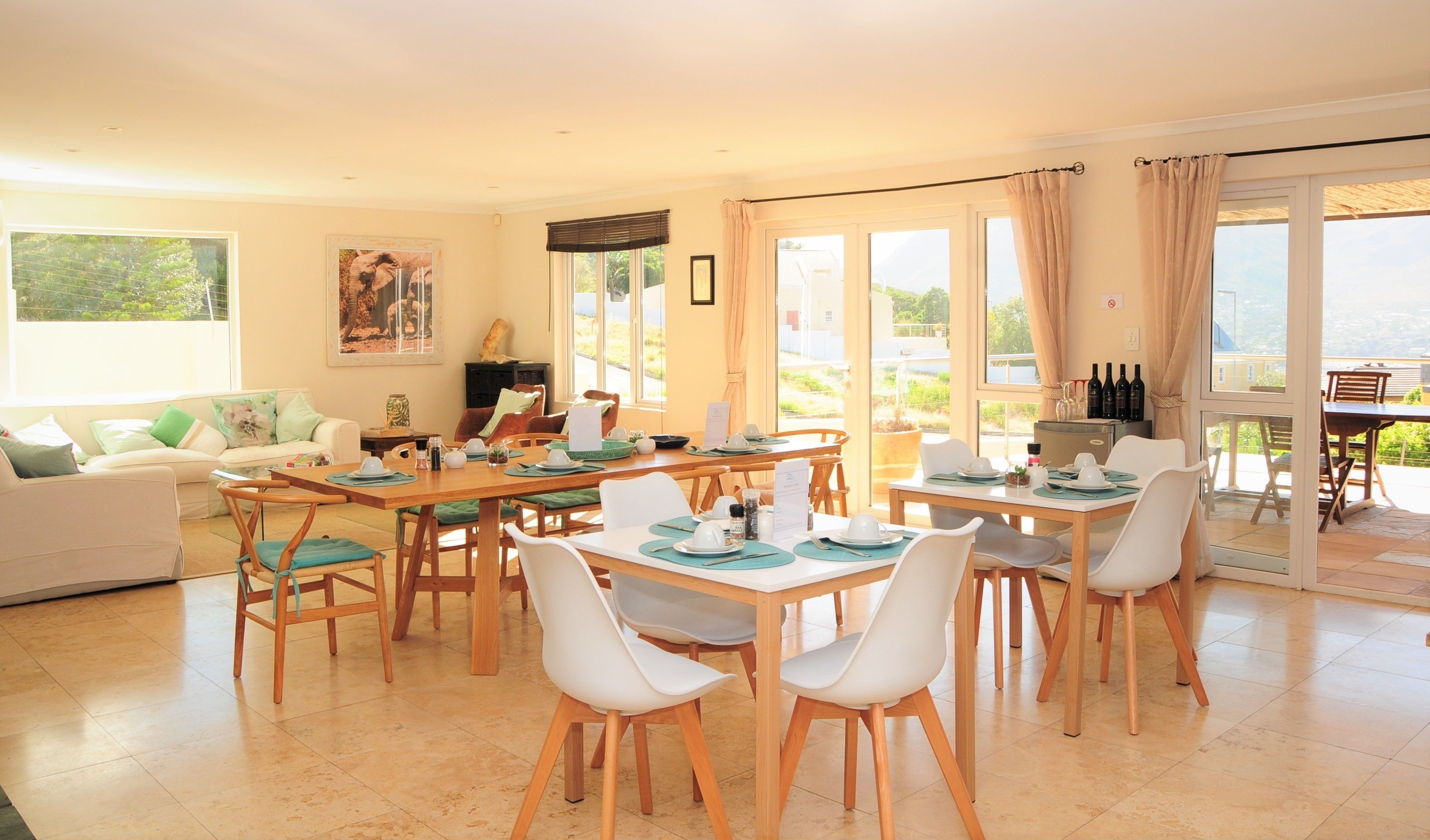 Un coin repas avec une table dressée pour quatre personnes, entourée de chaises blanches, dans une pièce lumineuse dotée de grandes fenêtres et offrant une vue sur la verdure à l’extérieur.