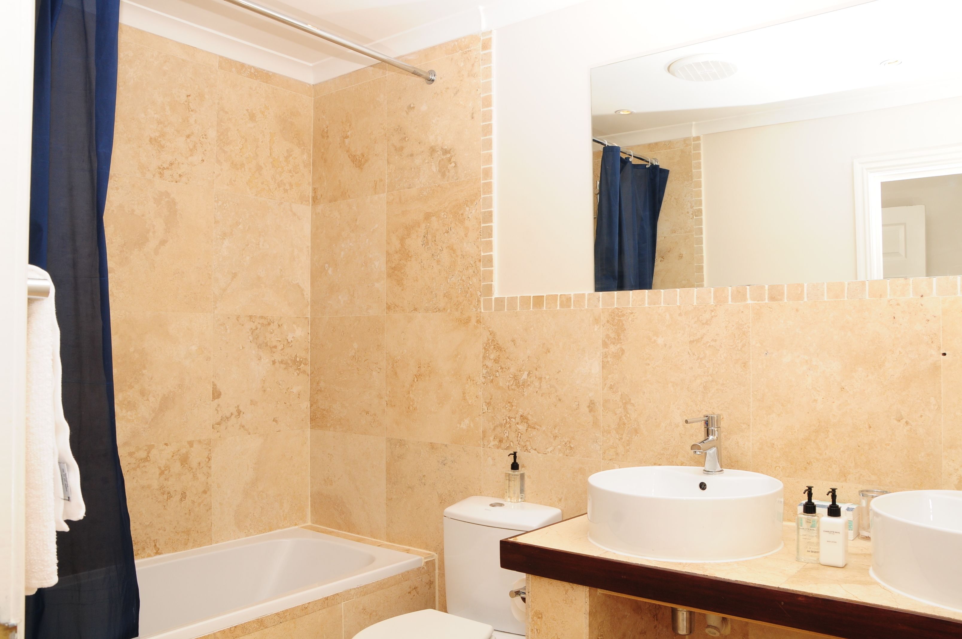 A bathroom with beige tiles, a bathtub, a toilet, and two sinks with mirrors.