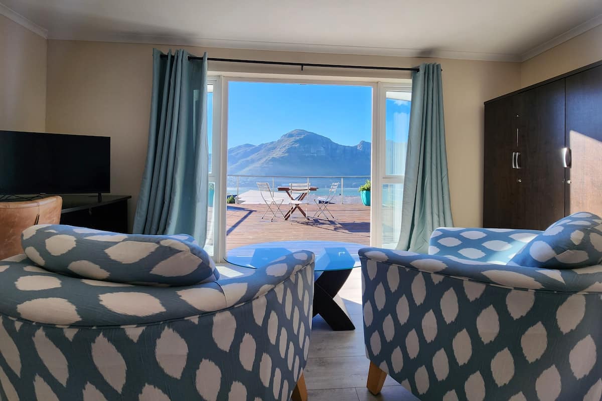 Ein Zimmer mit großem Fenster, einem Fernseher, zwei gemusterten Sesseln und einem Holztisch mit Stühlen davor.