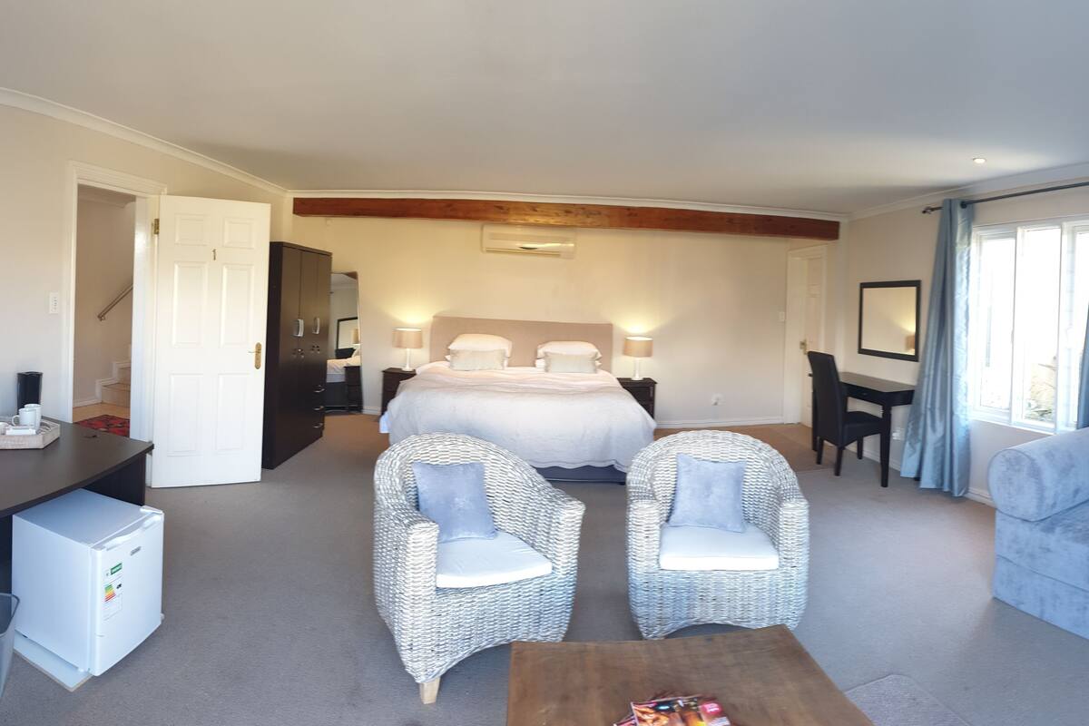 Ein Hotelzimmer mit einem Bett, zwei Korbstühlen, einem Schreibtisch, einem kleinen Kühlschrank, einem Holztisch und einem großen Fenster mit Vorhängen.