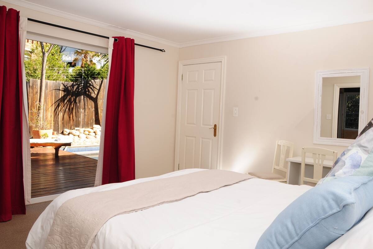 Ein Schlafzimmer mit Bett, Fenster, Tür und Blick auf einen Außenbereich mit Pool und roten Vorhängen.