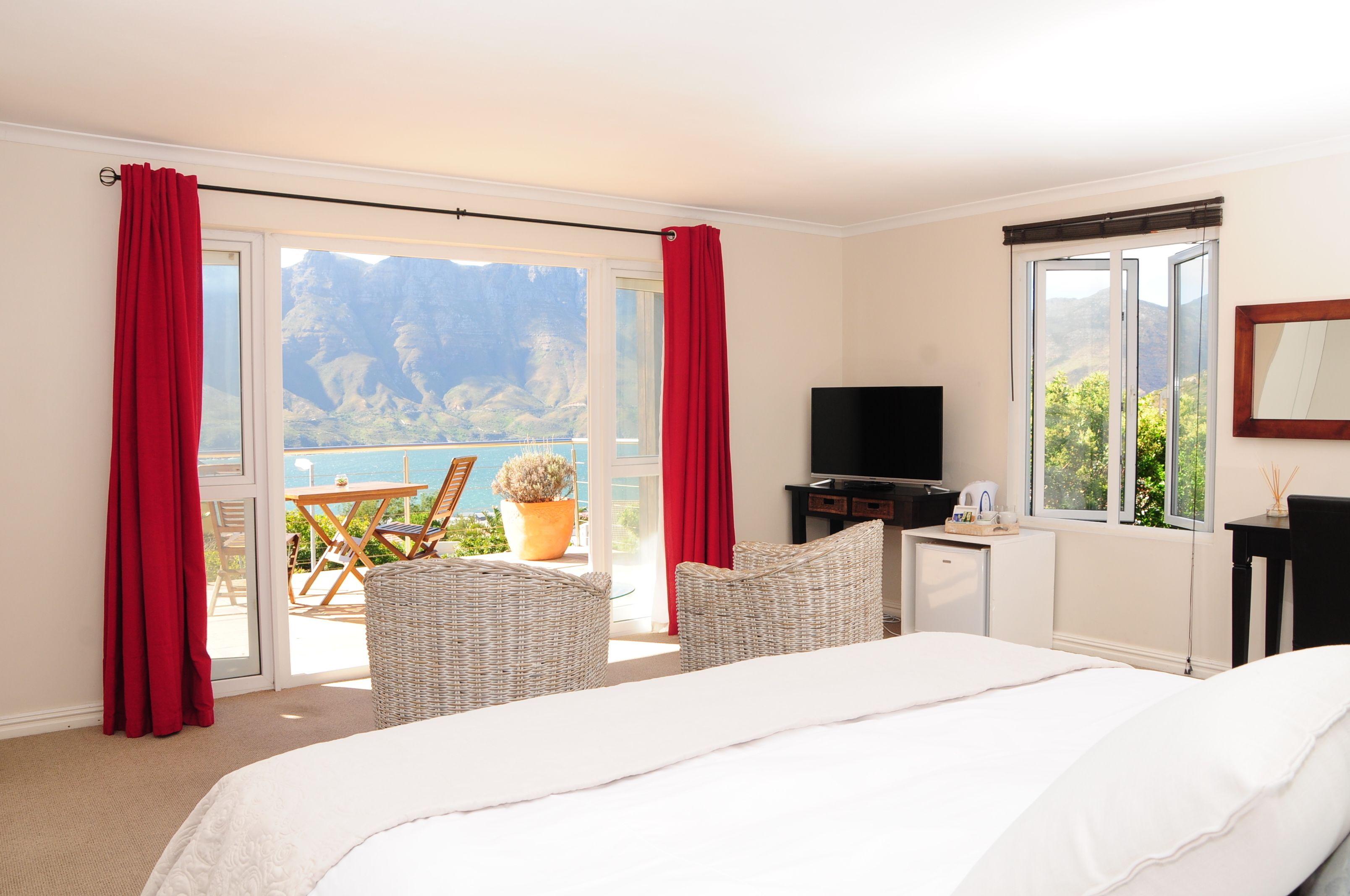 A hotel room with a bed, a TV, a small desk, wicker chairs, and a balcony with red curtains.