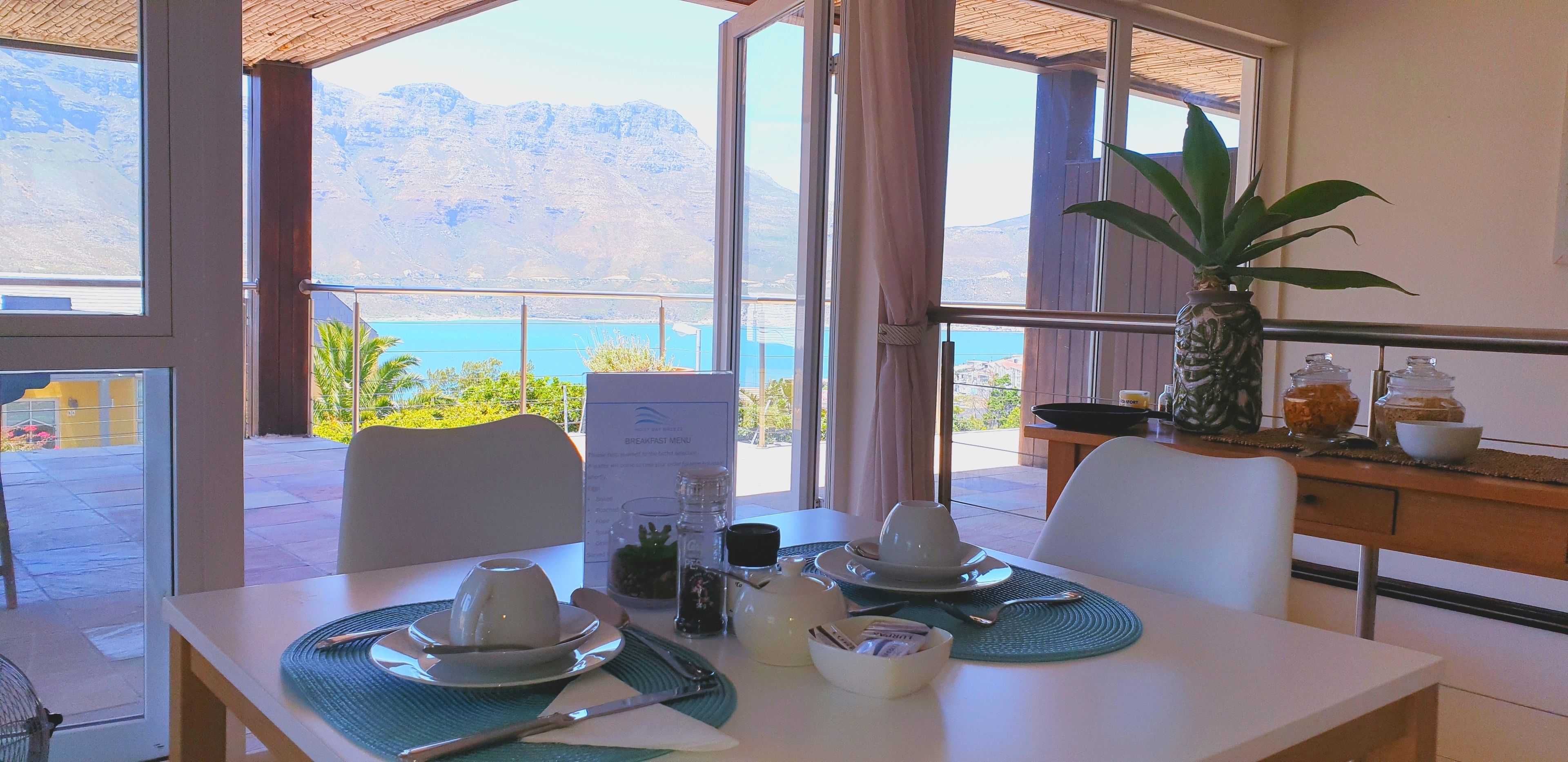 A breakfast table set with cups, a bowl, and utensils, overlooking a scenic mountain view and a body of water.