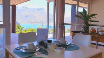 A breakfast table set with cups, a bowl, and utensils, overlooking a scenic mountain view and a body of water.