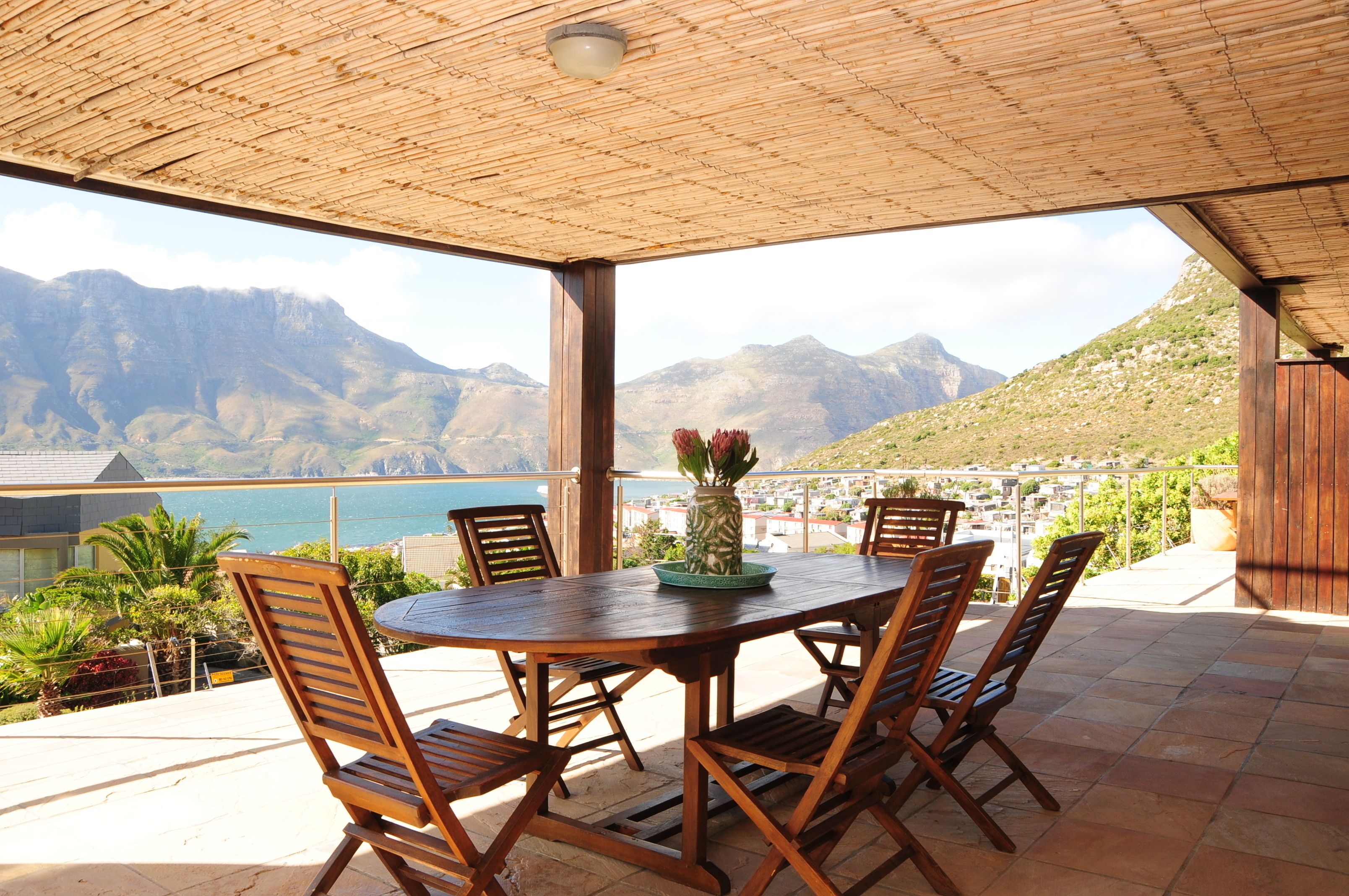 A covered outdoor dining area with a table and chairs, overlooking a mountainous landscape and a body of water.