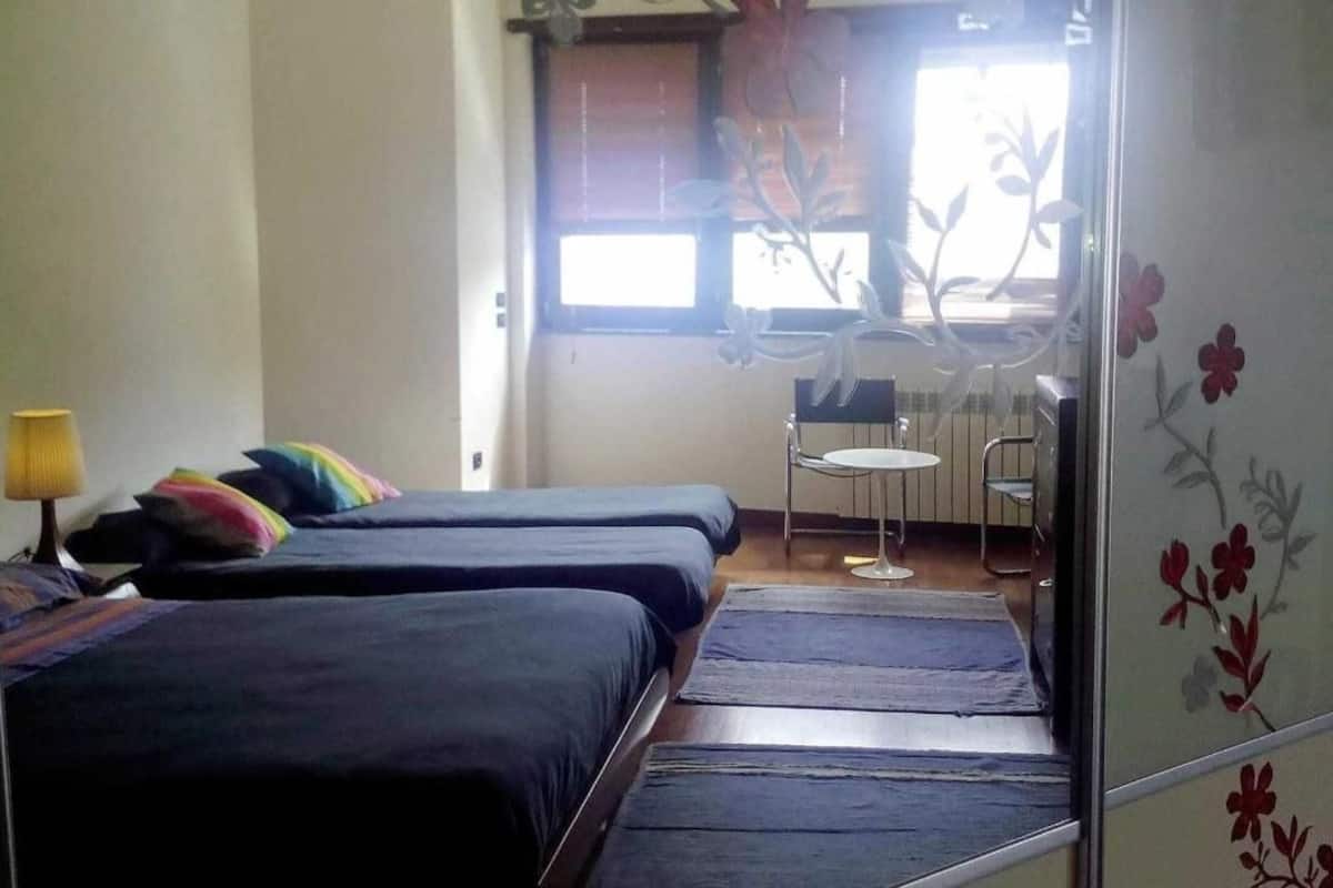 Ein Zimmer mit zwei Betten, einem kleinen Tisch und einem Fenster mit blumigen Vorhängen.
