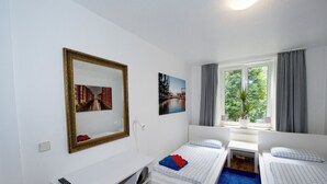 A hotel room with a bed, a desk, a chair, a mirror, and a window with curtains. - Basement Hamburg - Hostel (Hamburg)