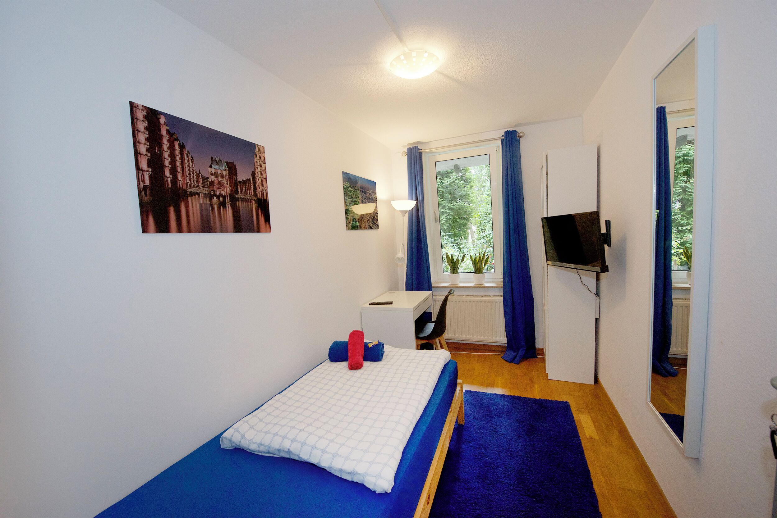 A bedroom with a bed, a desk, a chair, a TV, and a window with curtains.