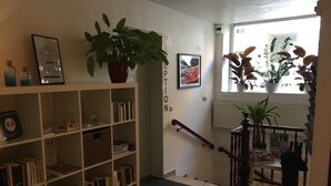 A library with bookshelves, a potted plant, a framed certificate, and a staircase with a handrail. - Basement Hamburg - Hostel (Hamburg)