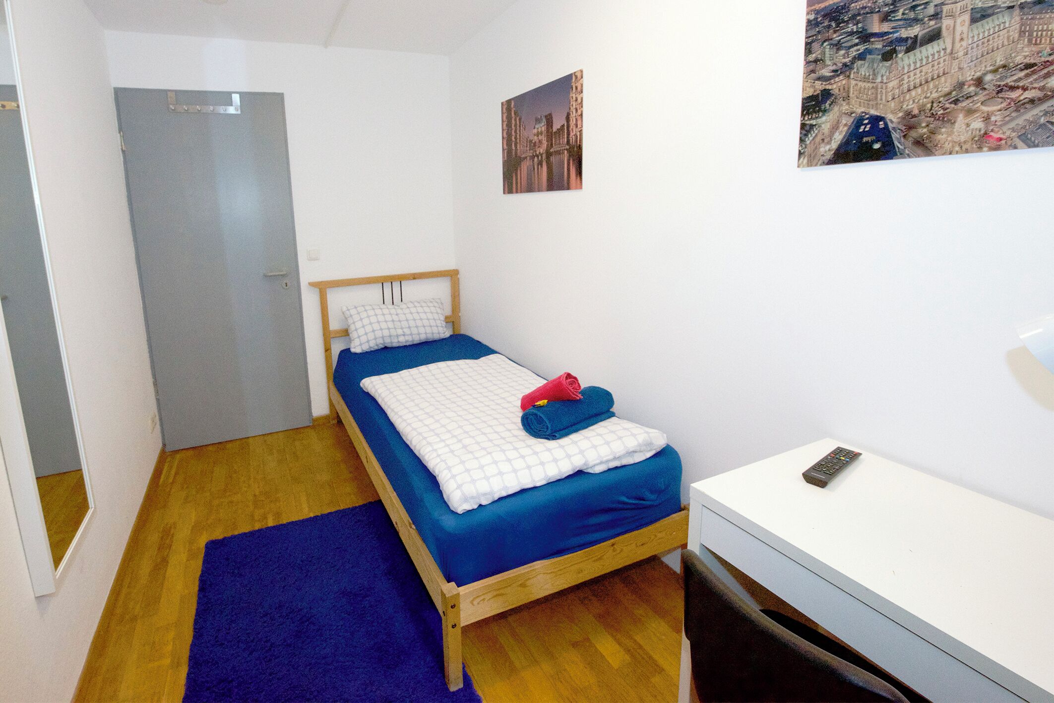 A single bed with a blue mattress and white bedding, a wooden headboard, a blue rug, a white desk, and a cityscape painting on the wall.
