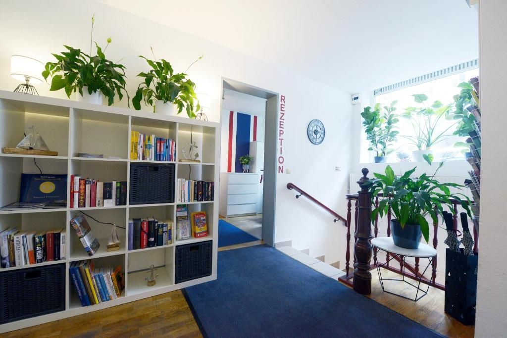 Zona de recepción con una estantería, alfombra azul y una escalera que lleva a otra habitación.