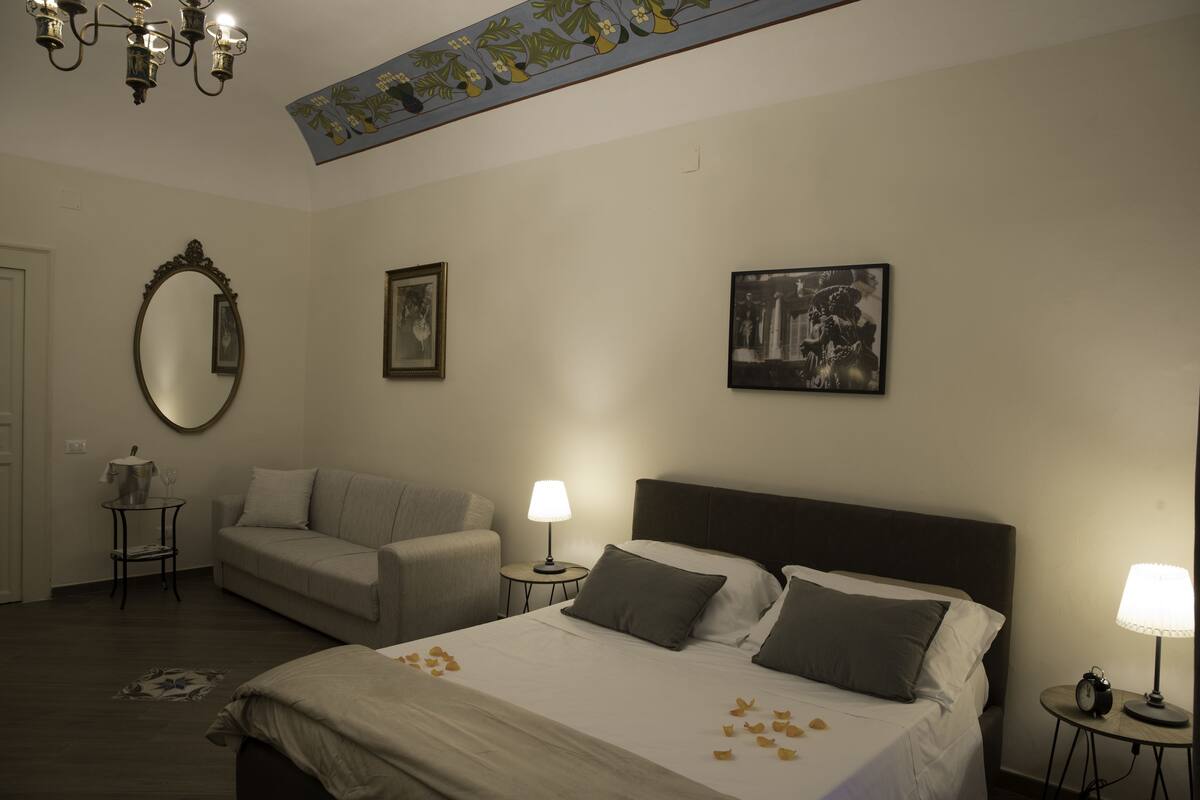 A hotel room with a bed, two bedside tables, a chair, a small table with a lamp, a mirror, and framed pictures on the wall.