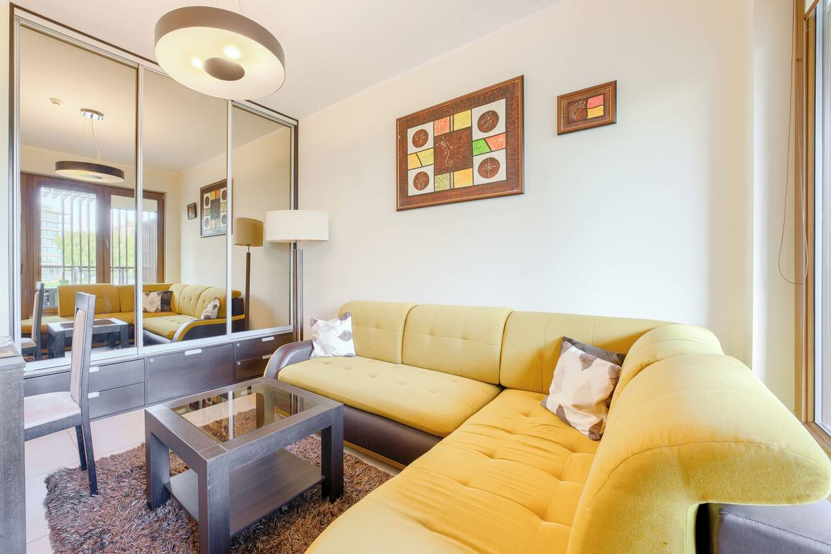 Ein modernes Wohnzimmer mit einem gelben Sofa, einem Glastisch und einem großen Spiegel, der den Raum widerspiegelt.