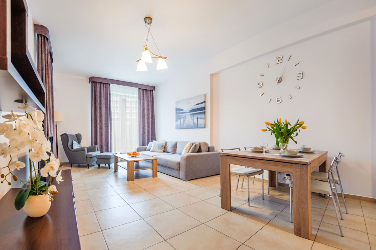 Ein modernes Wohnzimmer mit einer Couch, einem Couchtisch und einem Esstisch. Eine Chandelüster, ein Gemälde an der Wand und eine Uhr sind ebenfalls vorhanden.