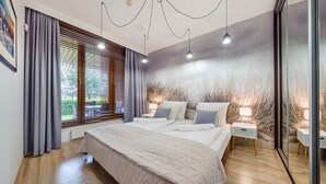 A modern bedroom with a large bed, a nightstand, a lamp, and a view of the outdoors through the window. - Sun & Snow - Olympic Park (Kolobrzeg)
