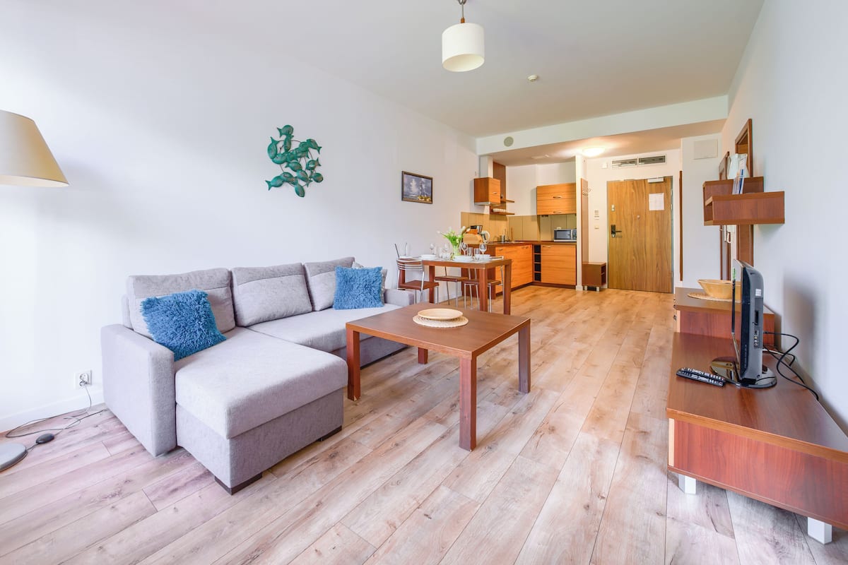 Ein modernes Wohnzimmer mit einem grauen Sofa, einem Holztisch und einer TV-Kommode. Im Hintergrund ist eine Küche zu sehen.