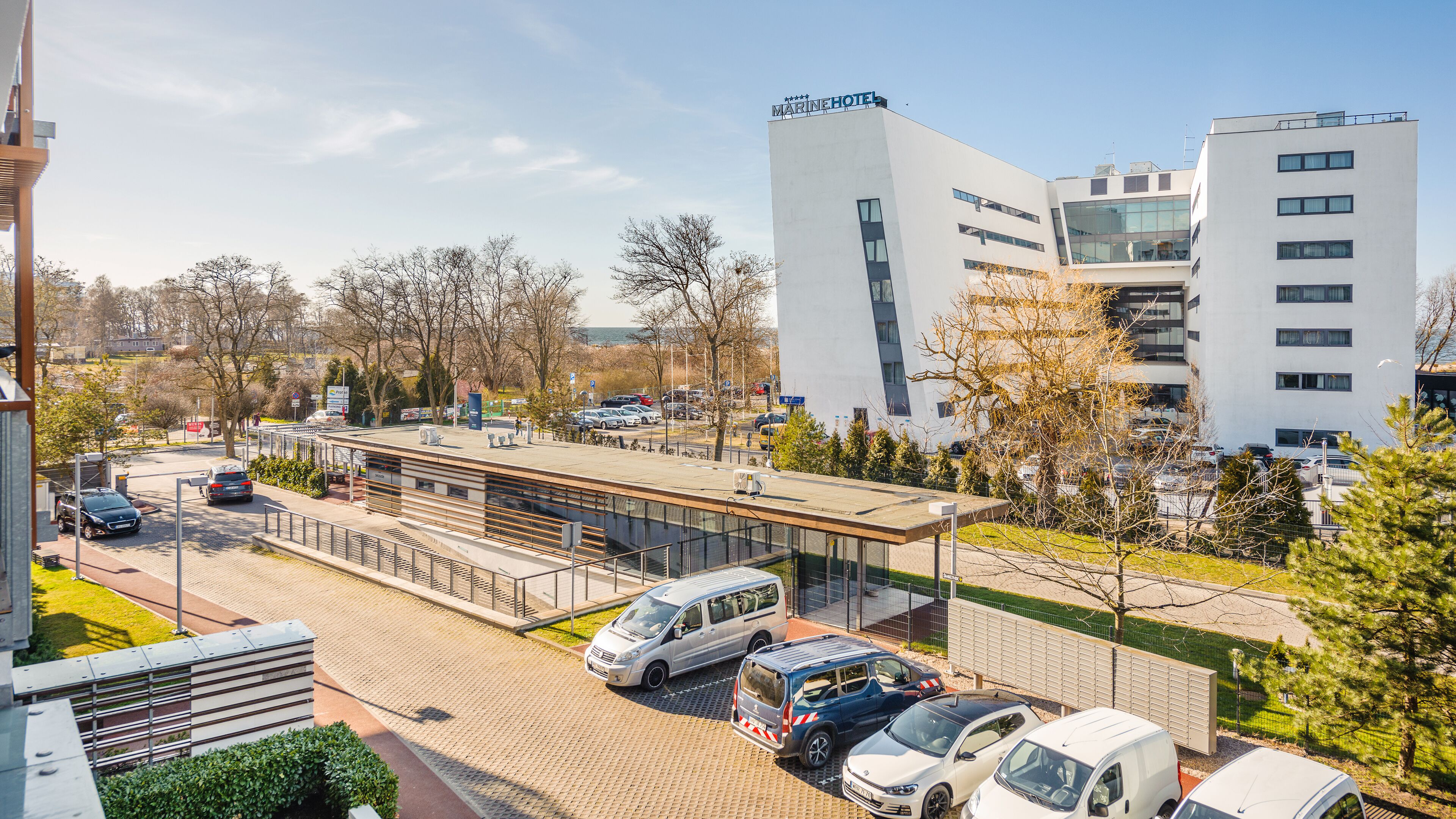 A modern building with a sign reading "MARINEHOTEL" and a parking area with several cars.
