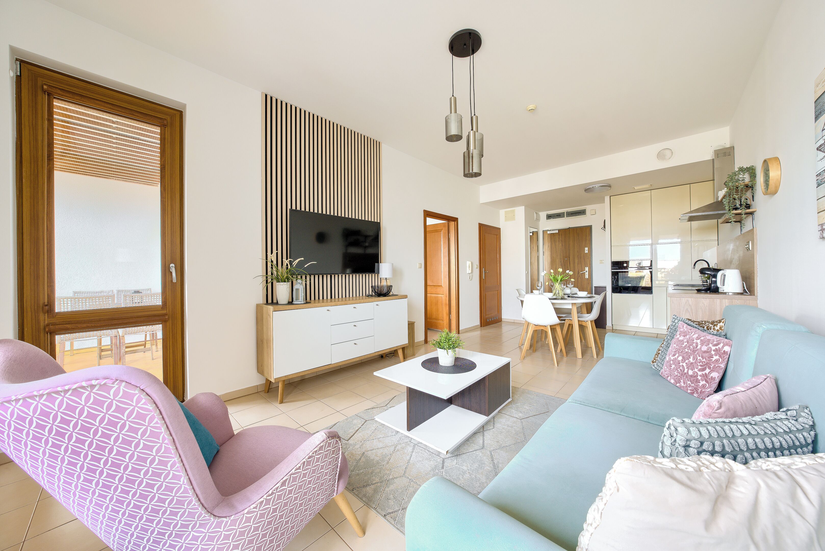 Una sala de estar moderna con un sofá, un sillón y una mesa de centro. Hay un televisor montado en la pared, un comedor con mesa y sillas, y una cocina al fondo.