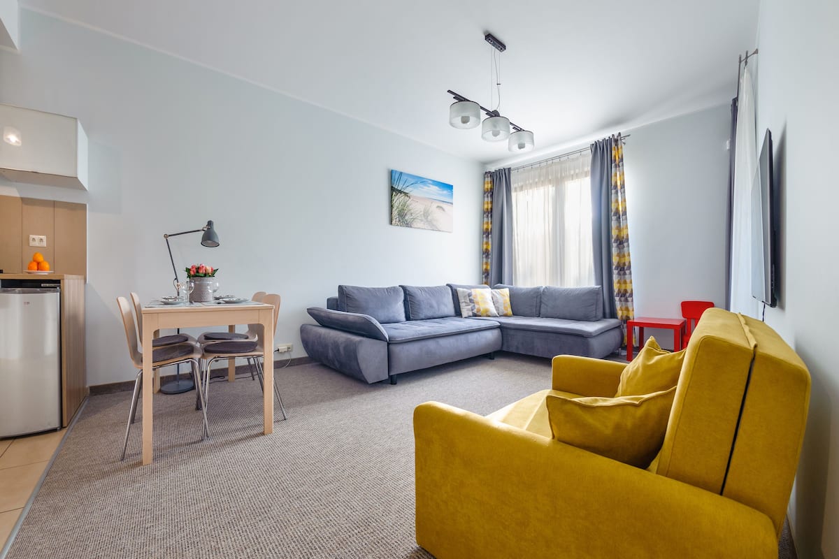 Ein modernes Wohnzimmer mit einer Couch, einem gelben Sessel, einem Esstisch mit Stühlen, einer Küchenzeile und einem großen Fenster mit Vorhängen.